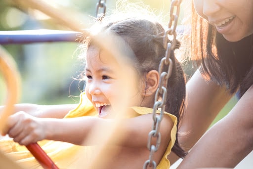 A girl smiles and laughs as her mom pushes her on a swing. 