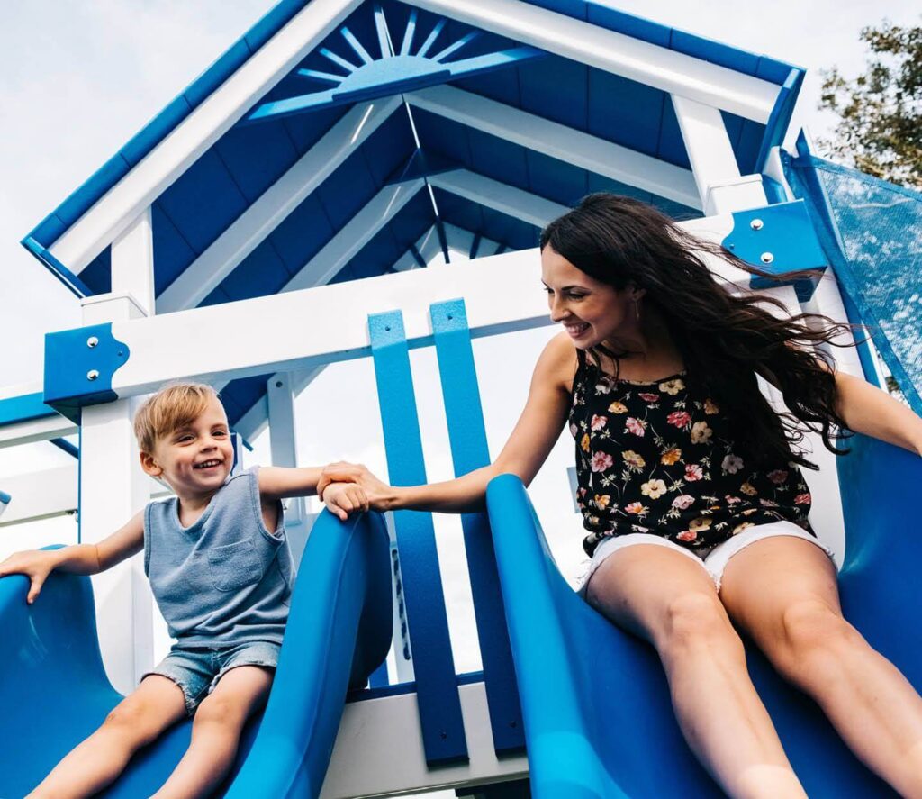 A mom and son holding hands, sliding down a set of double slides on a Swing Kingdom playset.